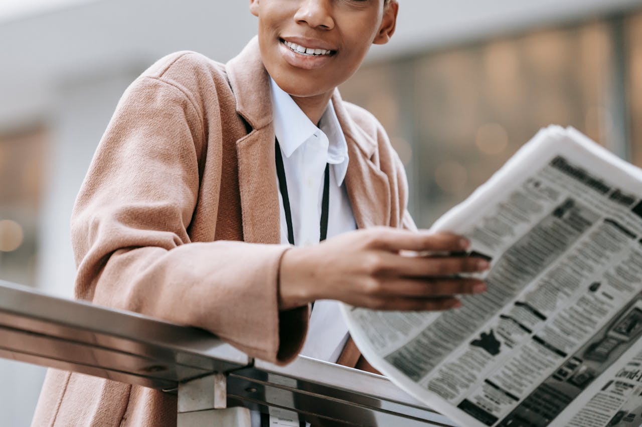 services-06 Crop happy African American businesswoman in formal clothes reading newspaper and looking away with smile while leaning on railing outside blurred modern building