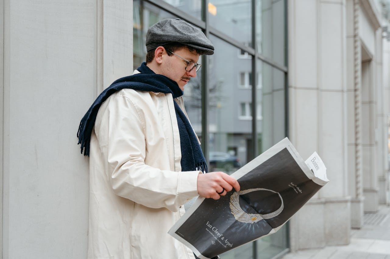Man wearing a flat cap and eyeglasses reads a newspaper outside an urban building.
