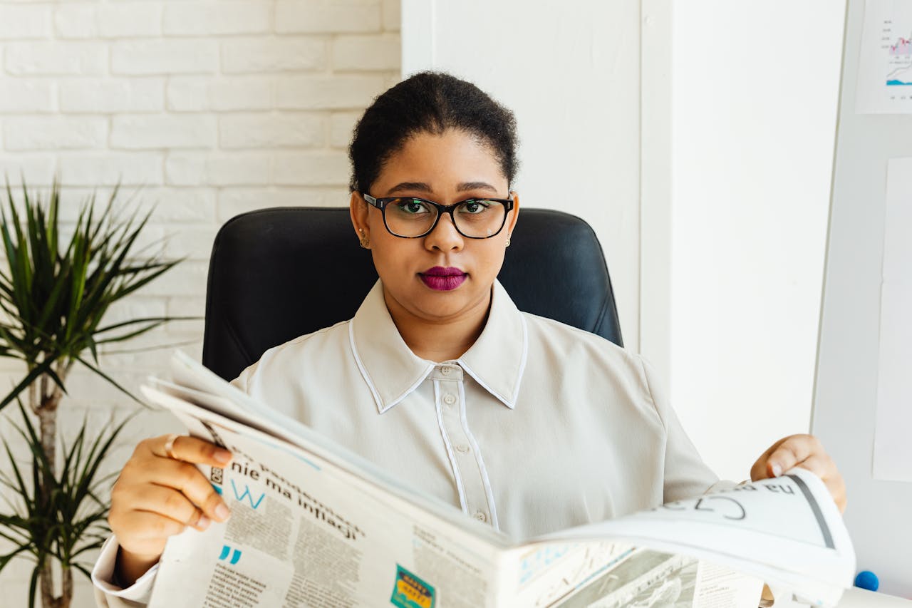Professional woman with glasses reading a newspaper at her office desk, surrounded by modern decor.
