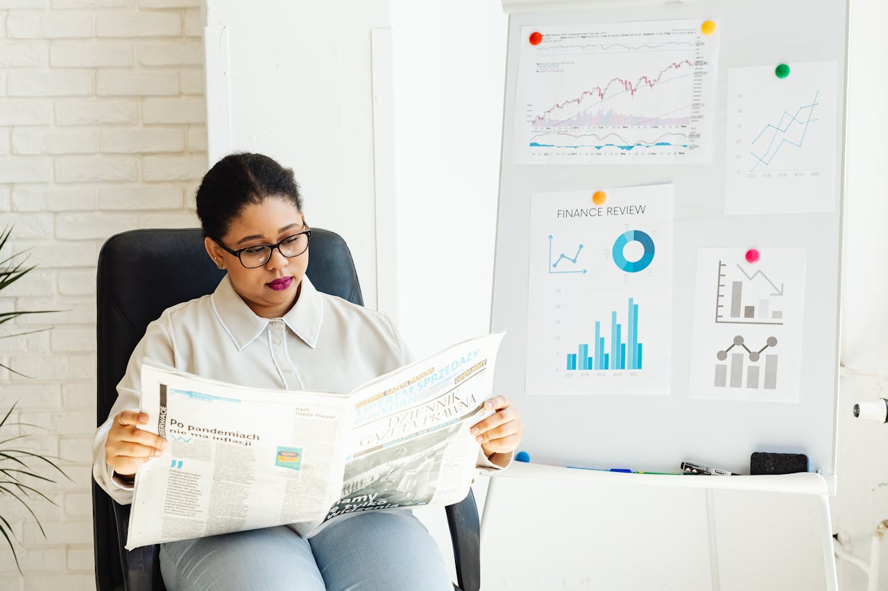 services-05 A woman reads a newspaper with stock charts displayed in an office setting.