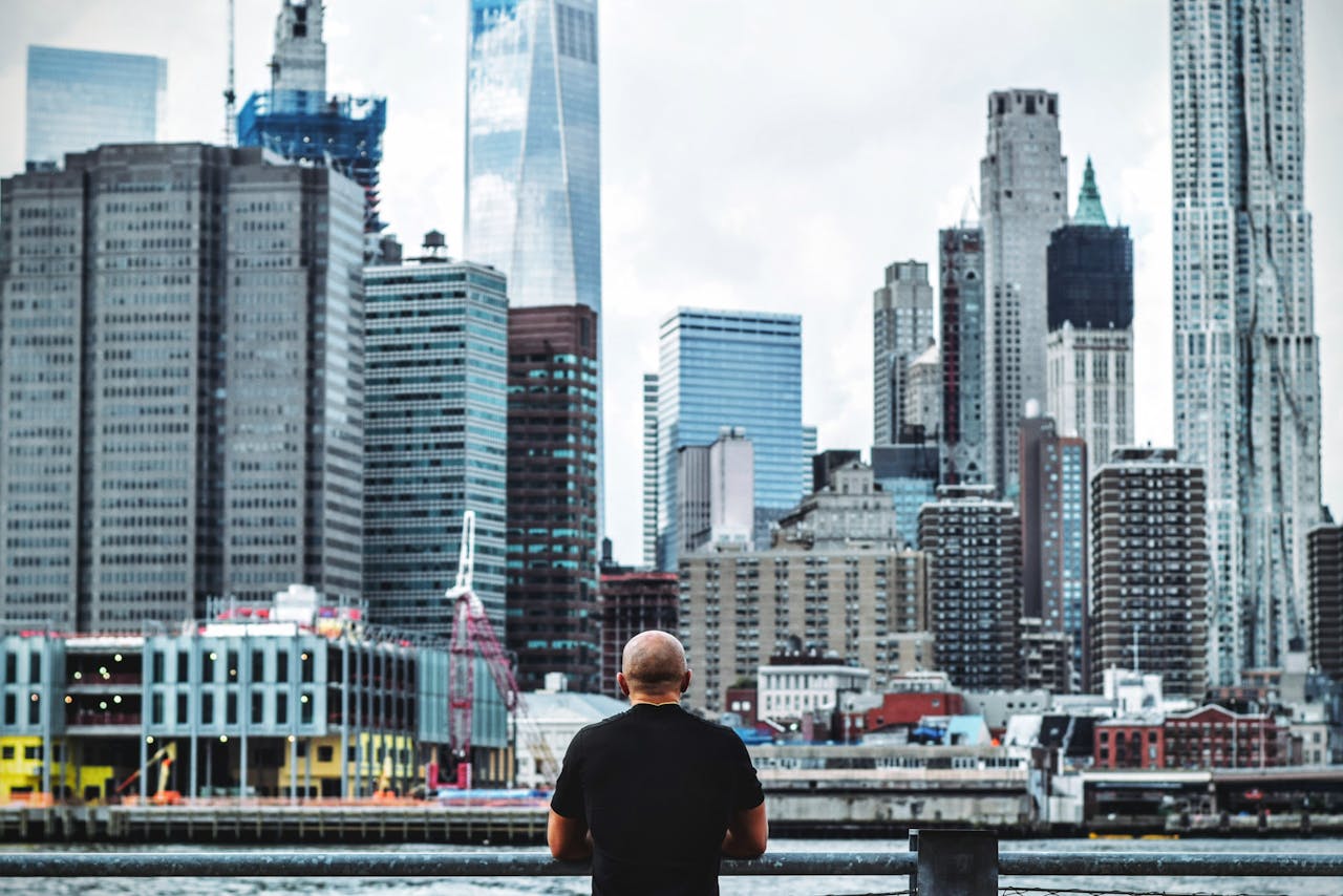 Home A person stands in Brooklyn, gazing at the panoramic Manhattan skyline, featuring skyscrapers.