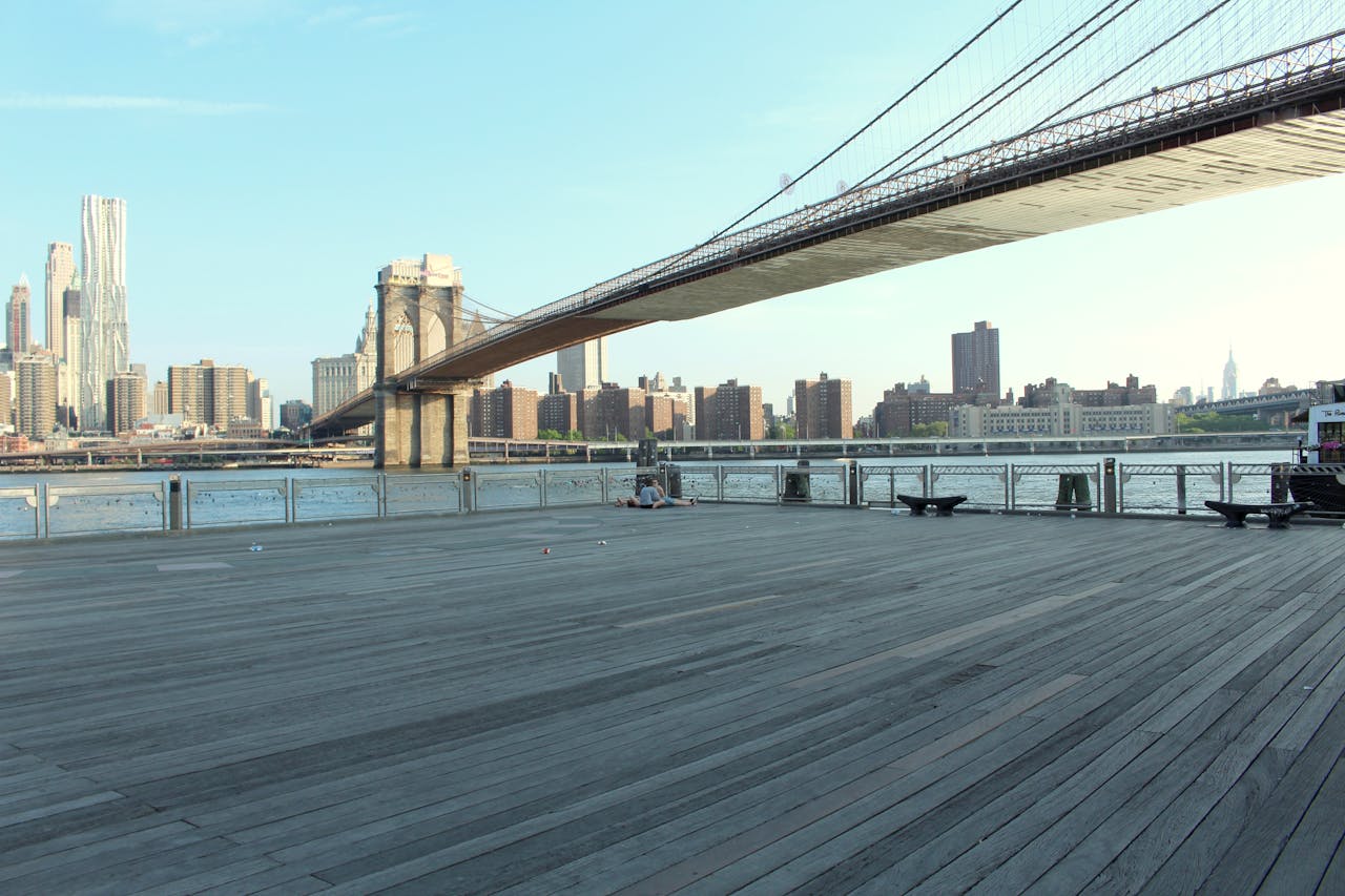 Home Scenic view of the Brooklyn Bridge and Manhattan skyline from a waterfront boardwalk in New York City.
