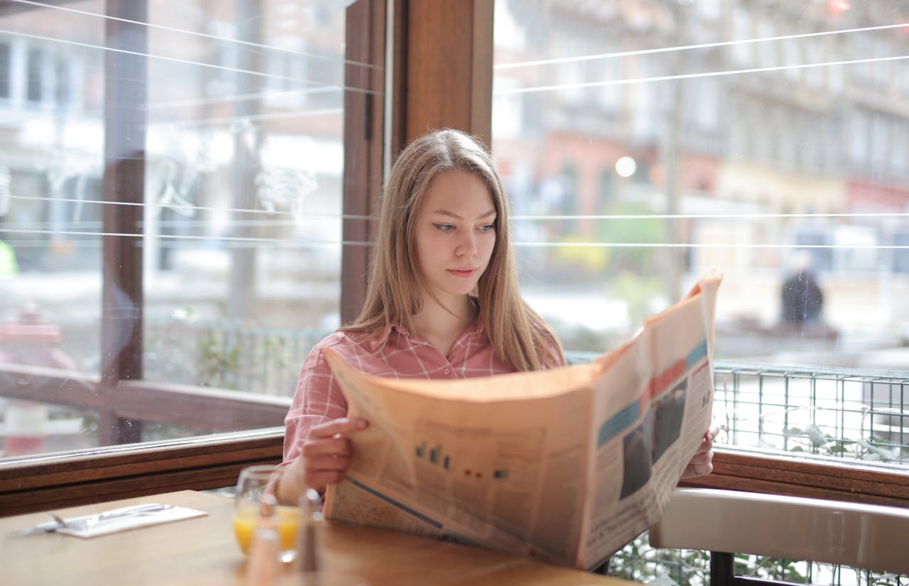 services-04 A young woman enjoying a newspaper at a cafe table, immersed in reading.