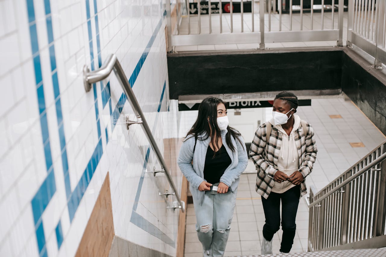 services-05 Two young women in masks ascend stairs in an urban subway station, embracing new norms.