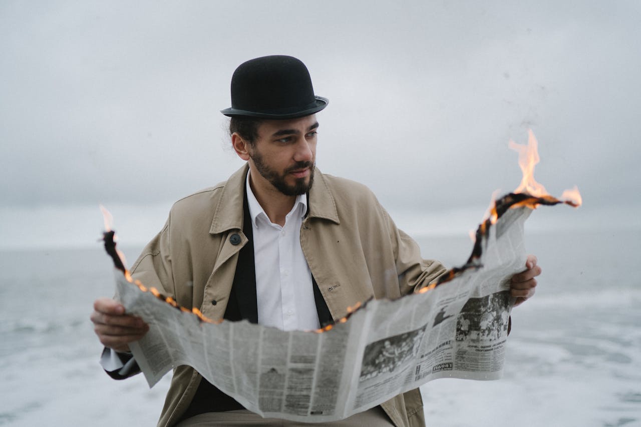 hero-img A man in a bowler hat and coat reads a burning newspaper by the ocean on a cloudy day.